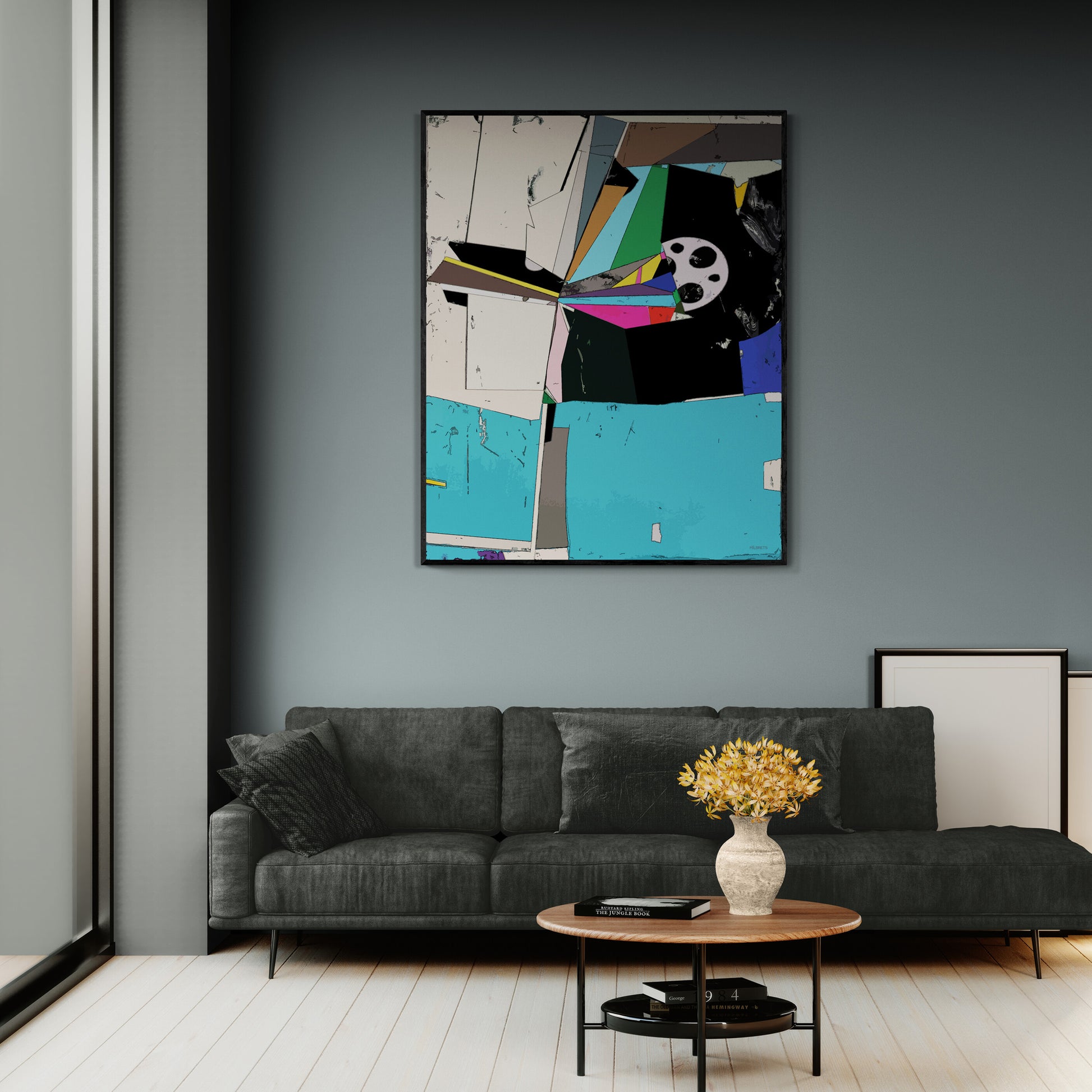 A framed abstract wall art print with geometric shapes and bright color blocks hangs on a dark gray wall above a modern dark gray sofa in a styled living room.