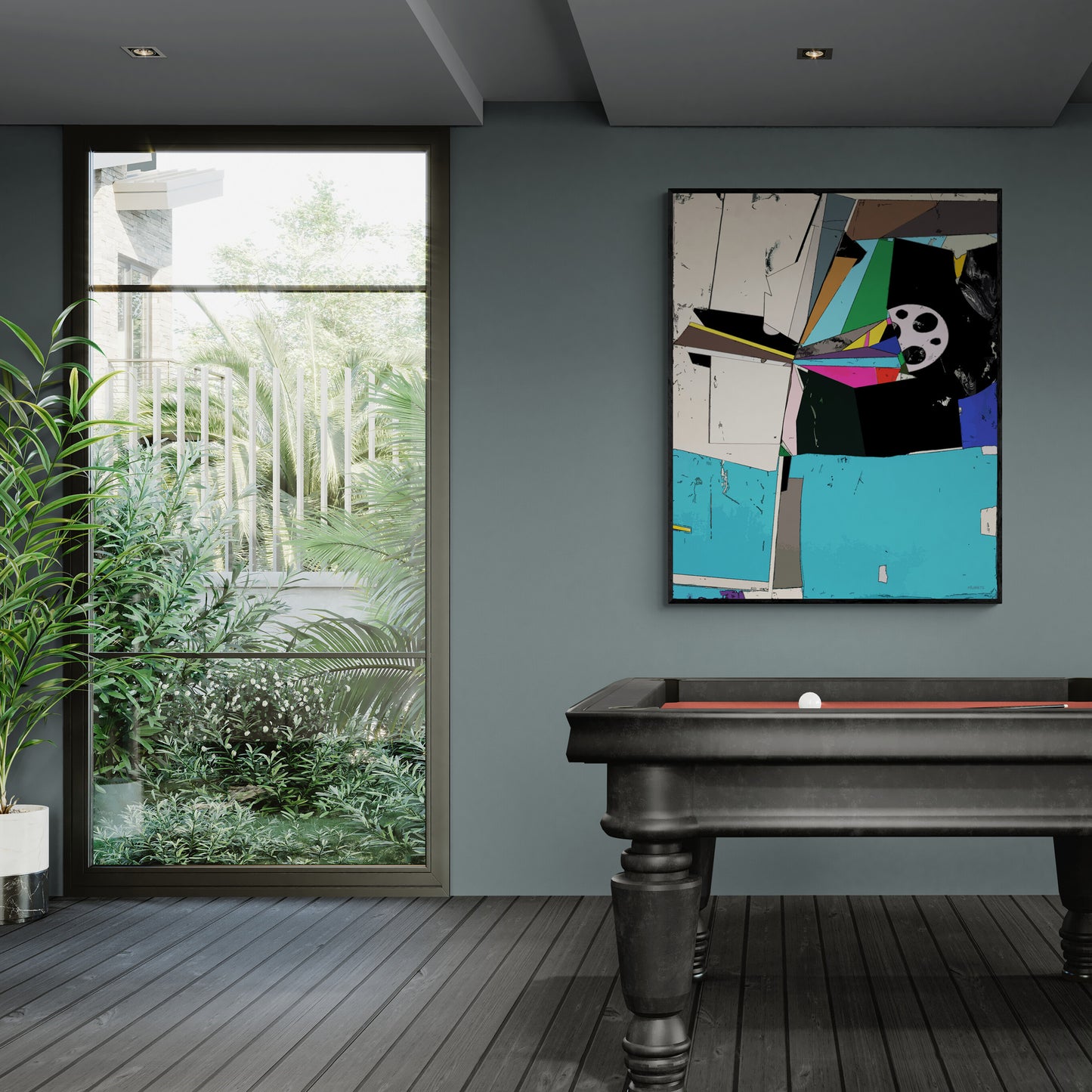 A framed abstract geometric wall art print with bold blocks of turquoise, black, white, and pink, hanging on a dark gray wall above a pool table in a modern room with a large window overlooking greenery.