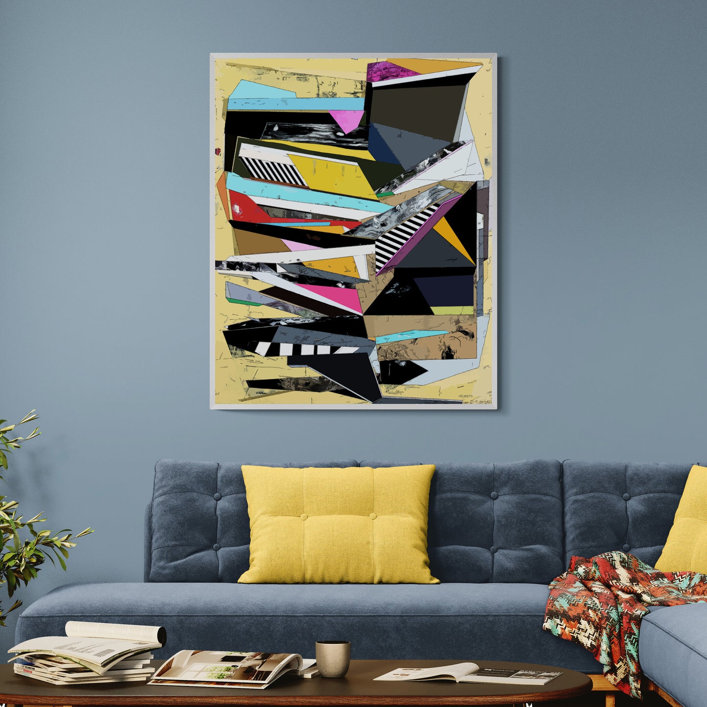 A framed abstract geometric wall art print with bold black, white, yellow, pink, blue, and gray shapes on a beige background, displayed above a blue sofa with yellow pillows in a living room setting.