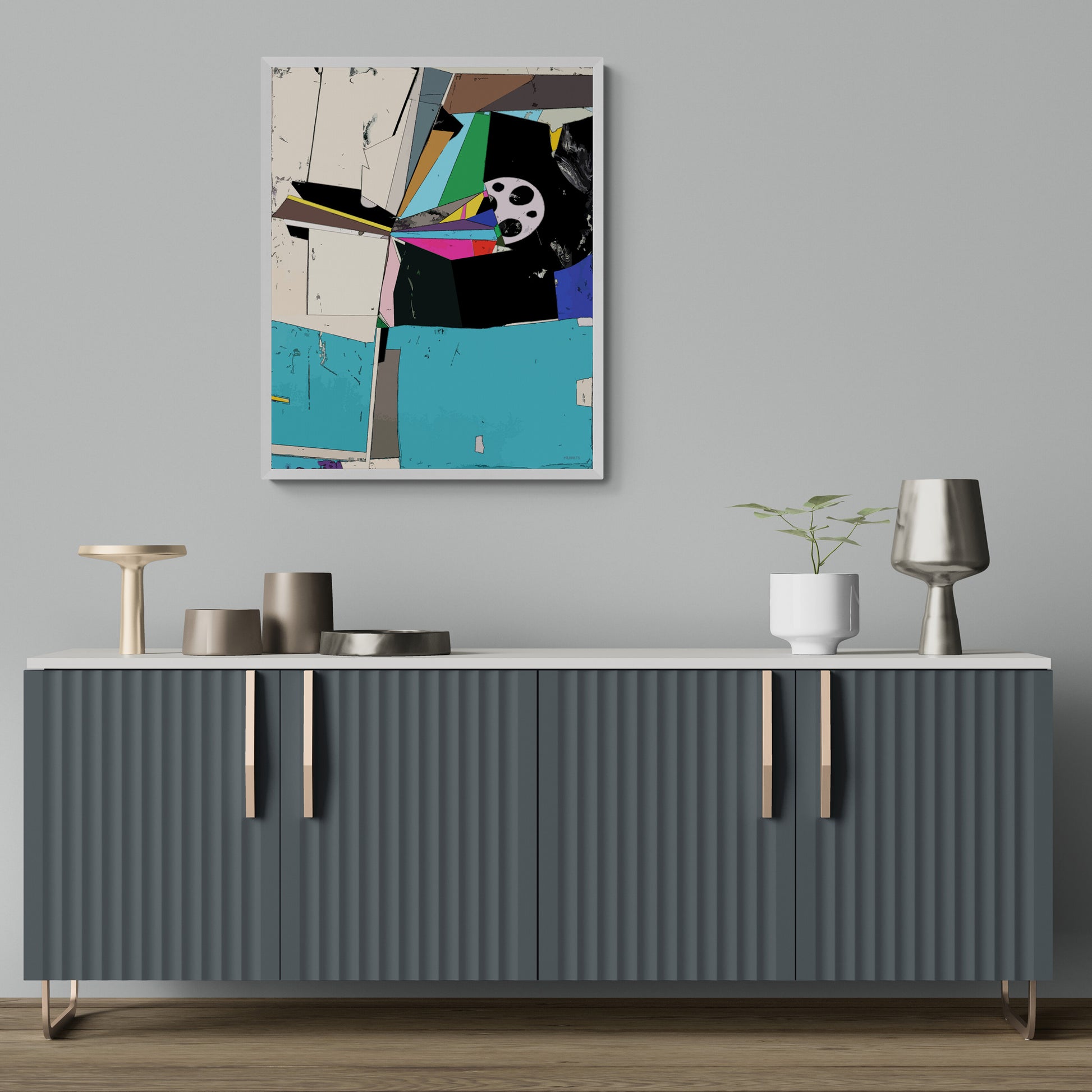 An abstract geometric wall art print with a white frame hangs on a light gray wall above a modern gray ribbed sideboard with decorative objects and a small plant.